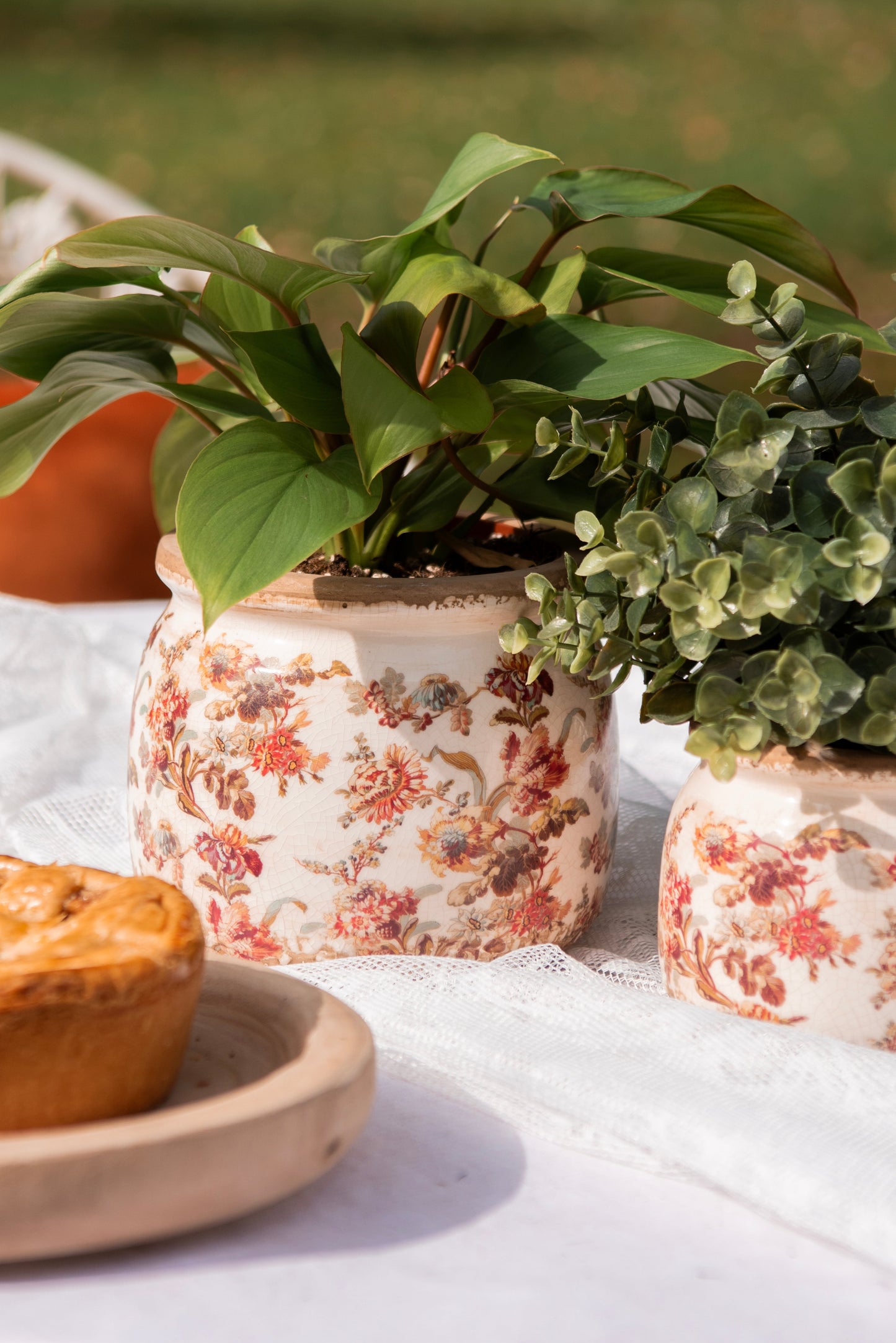 Une table en extérieur ornée de pots de fleurs en céramique au motif floral vintage, contenant des plantes vertes luxuriantes. Un petit plat en terre cuite accueille une tourte dorée, apportant une touche gourmande à cette scène chaleureuse et élégante. En arrière-plan, un jardin verdoyant et un coussin orange sur une chaise en fer forgé complètent cette ambiance douce et bucolique.